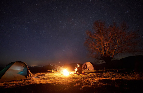 晚上在山上露营。 游客坐在草地上的两个照明帐篷之间，享受着明亮燃烧的篝火。 美丽的深蓝色星空和背景上的大树