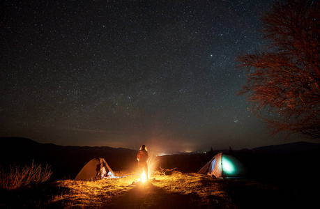 晚上在山上露营。 女游客站在两个帐篷之间的剪影，在大树附近燃烧的篝火上，享受着深邃的黑暗天空，有很多明亮的星星。 旅游和旅游概念