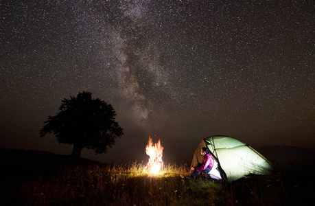 在山上露营的夜晚。 年轻的徒步旅行者女人坐在明亮的帐篷入口，在美丽的深黑的星空和银河下享受着明亮的燃烧的篝火。 旅游和旅游概念