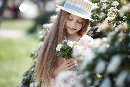 肖像美丽的快乐女孩享受气味在一个开花的春天盛开的花园。 明亮时尚的微笑女孩在春天的花园里开着美丽的灌木树和白花。 景观。 戴帽子