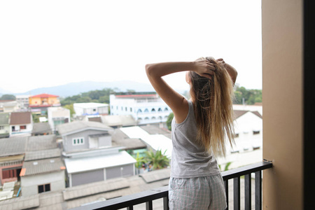 穿着夏季睡衣的年轻金发女孩站在阳台上, 看着举起手的建筑物