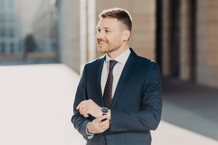 侧面拍摄的快乐男经理穿着正式西装,保持手在手表上,愉快地看着远方