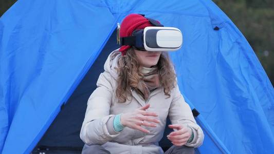 迷人的年轻女子游客在一个红色的帽子坐在旅游帐篷和享受虚拟现实头盔