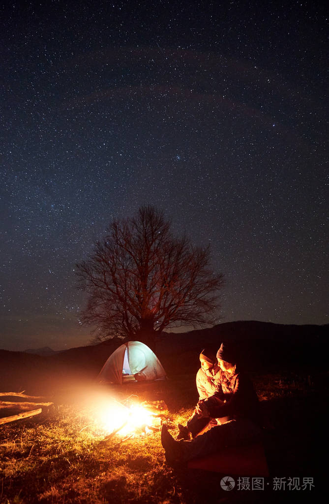 在山上露营的夜晚。 情侣游客男孩和女孩在星空下燃烧的篝火旁休息。 照亮帐篷大树和远处的山丘背景。 旅游和旅游概念。