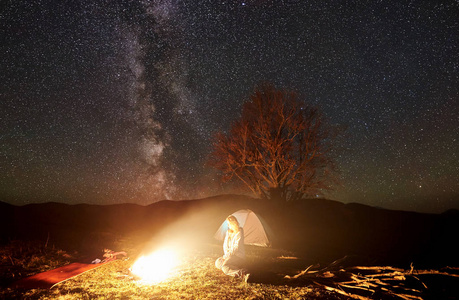 在山上露营的夜晚。 女游客休息在燃烧的篝火下星空和银河。 背景中大树的照明帐篷和剪影。 旅游和户外活动概念