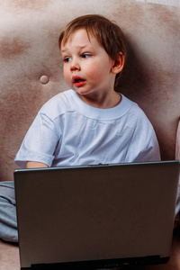 穿着白色T恤的小男孩坐在椅子上，沙发上有一台笔记本电脑。看着相机。有趣的脸。集中注意力。