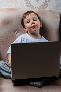 穿着白色T恤的小男孩坐在椅子上，沙发上有一台笔记本电脑。看着相机。有趣的脸。集中注意力。