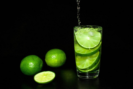 冷脱毒水与冰和新鲜成熟切片绿色石灰在黑色背景。 夏季清爽和饮料的概念。 冷脱毒水与冰和新鲜成熟切片绿色石灰在黑色背景。