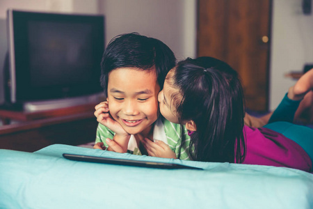 快乐的亚洲儿童微笑和使用数字平板电脑。 姐姐亲吻她哥哥的脸颊，俯卧在床上。 爱兄弟姐妹的概念。 老式电影过滤效果色调。 电影风格