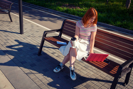 一个年轻的红头发女人穿着一条白色浪漫的裙子，一个粉红色的上衣和一双蓝色的运动鞋，在她旁边的城市公园里喝咖啡，坐在一张长凳上，是一
