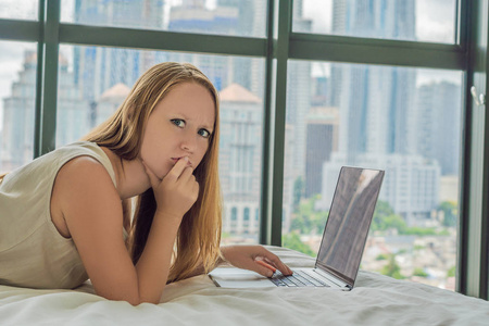 年轻女子正在他的床上的笔记本电脑上的背景下, 俯瞰摩天大楼的全景窗口。自由职业者, 远程工作, 在家工作