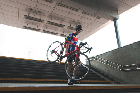 穿着运动服的自行车手里拿着一辆自行车走下地下通道的楼梯。在地铁里骑自行车。骑着自行车环游城市。