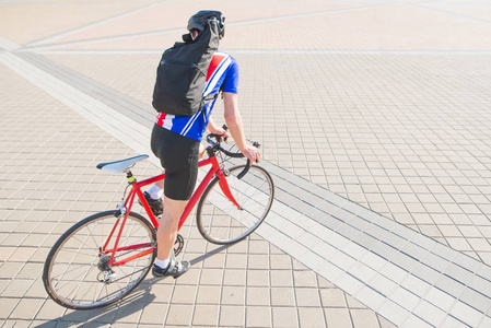 带着背包的Yclist骑着一辆红色自行车在一个有人行道的广场上。 一个穿运动服和头盔的人在城里骑自行车。