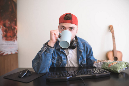 一个玩家坐在家里的电脑上，拿着杯子和零食的肖像。 戴着帽子和耳机的年轻人从杯子里喝东西，吃零食，在家里电脑上玩电子游戏。 游戏者