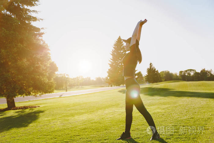 年轻的运动女孩穿着黑色制服帽做运动锻炼热身伸展然后在阳光明媚的