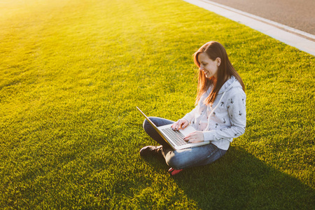 穿休闲服装的年轻学生女性。 女人坐在草地上，在户外绿草阳光草坪上的城市公园里，在笔记本电脑电脑上工作。 移动办公室。 自由职业商