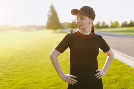 年轻的运动女孩穿着黑色制服帽，做运动锻炼，热身伸展，然后在阳光明媚的夏日在高尔夫球场公园户外的绿色草坪上跑步。 健康的生活方式