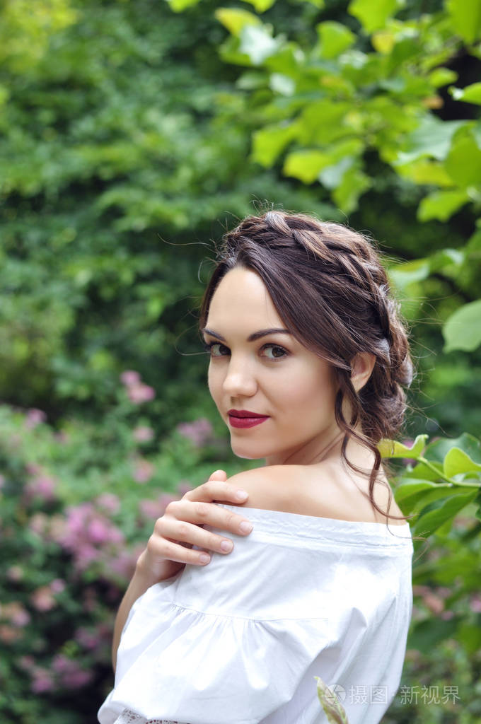 年轻漂亮的深色头发的女人穿着彩色连衣裙敞着肩膀在明亮的绿色盛开的