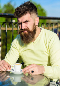 有胡子的男人用咖啡杯, 喝咖啡。咖啡打破概念。胡子和胡子的人在严密的面孔, 阳台篱芭在背景, 弥散。长胡子的人看起来严厉和严肃