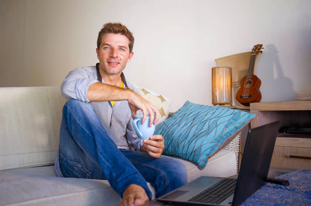 年轻有魅力和成功的男人，在家客厅工作，坐在笔记本电脑上的沙发网络上，喝着咖啡，在自由职业者和独立的工作概念中快乐