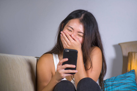 年轻甜美快乐漂亮的亚洲女孩在手机上使用互联网社交媒体应用程序，笑得开心，在家玩沙发沙发，在线交流生活方式概念