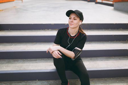 年轻的运动美丽的女孩，戴着黑色制服帽，戴着耳机，听着音乐，在户外城市公园的楼梯上跑步训练前后休息和坐着。 健康的生活方式