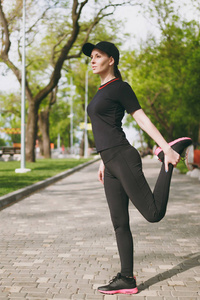 年轻的运动，美丽的黑发女孩，穿着黑色制服帽，做运动，伸展运动，热身，然后跑步或训练站在城市公园户外。 健康的生活方式