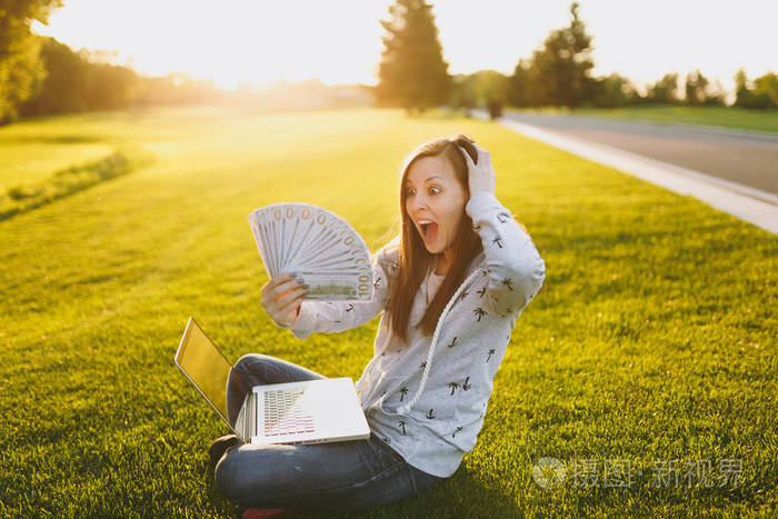 女性持有一捆美元现金。 女人坐在草地上，在户外绿草阳光草坪上的城市公园里，在笔记本电脑电脑上工作。 移动办公室。 自由职业商业概