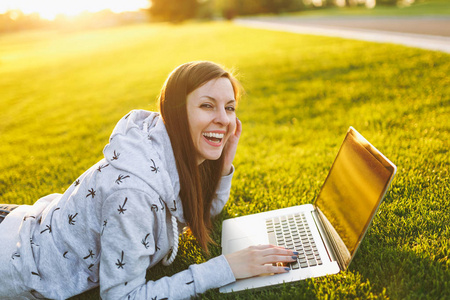 年轻聪明的女学生穿休闲服装。 女人躺在草地上，在户外绿草阳光草坪上的城市公园里，在笔记本电脑电脑上工作。 移动办公室。 自由职业