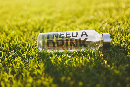 带有新鲜干净透明水的瓶子，上面有文字铭文，我需要在阳光草坪上的绿草上喝。 自然纹理绿色背景壁纸。 足球场或运动概念设计