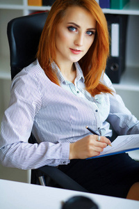 生意。 严肃女人的肖像。 商务女性红头发，坐在大光办公室的桌子旁，手里拿着一个文件夹，写字。 穿着白色衬衫和黑色裙子的女孩。