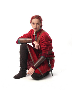 穿着幻想中世纪服装的红头发女孩的全长肖像，坐在演播室背景上的姿势。