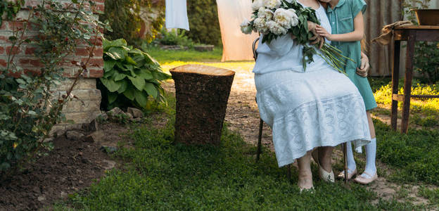 祖母一位穿着白色老式衣服的老妇人坐着拥抱她的孙女，一个穿着绿色连衣裙的女孩，另一只手拿着花园里一束白色牡丹花。 会议和祝贺礼物
