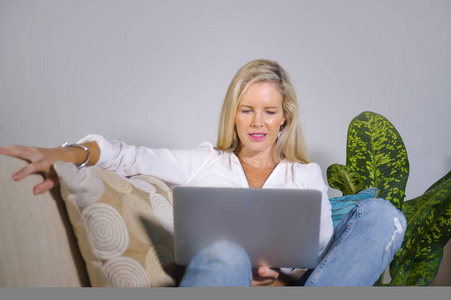 40岁出头的漂亮快乐优雅的金发女人在家庭客厅里放松，在笔记本电脑上上网，在现代公寓的沙发上舒适地工作，生活方式理念