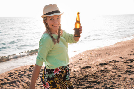 快乐的年轻女人戴着金色的辫子和草帽，在天堂海滩上喝了一瓶啤酒。 幸福概念
