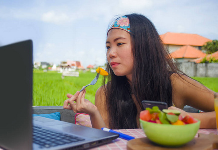 年轻有魅力快乐的亚洲韩国数字游牧民在户外工作，轻松地用笔记本电脑吃健康沙拉，在成功的工作中担任在线社交媒体旅游博主