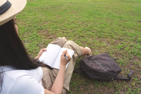 年轻的女人戴着帽子在笔记本上写着铅笔在户外公园的草地上。 大学女孩穿休闲白色衬衫棕色裤子和背包坐在花园里。 写作理念和教育理念。