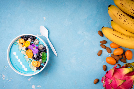 蓝色冰沙碗，有水果，浆果，坚果和花。热带健康冰沙甜点。健康饮食，素食，饮食理念..上景。平躺式