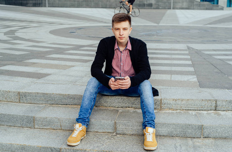 时尚的年轻人穿着格子衬衫和牛仔裤坐在楼梯上，在户外使用智能手机全身。 提供智能手机免费空间的学生