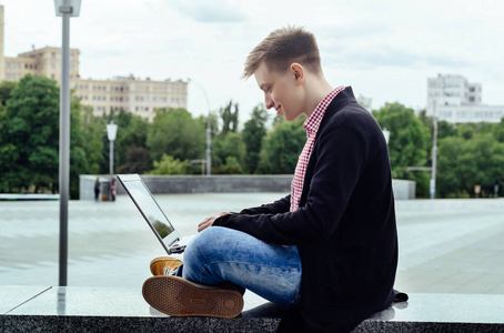 时尚的年轻人穿着夹克和牛仔裤，坐在笔记本电脑户外侧视图上打字。 技术和通信概念