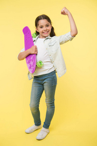 我可以骑它。小女孩快乐地拿着便士板。女孩快乐的脸扛着便士板黄色的背景。孩子学会了骑便士板。孩子喜欢塑料滑板显示力量手势。女孩力量