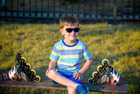 穿着滚筒刀片的小帅哥坐在夏天公园的长凳上。 快乐微笑运动的孩子戴着安全帽。 积极休闲暑假时间与家人。