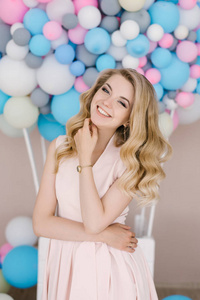 一个美丽的年轻女孩的肖像，卷发金发。 站在白色和蓝色气球的背景下穿着一件浅色的衣服。 精致的头发和妆容