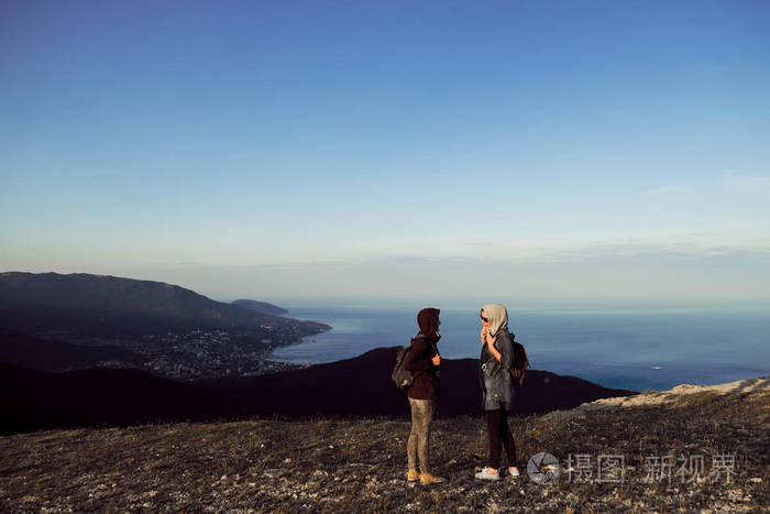 两个女孩旅行者背对着山顶边的镜头站着