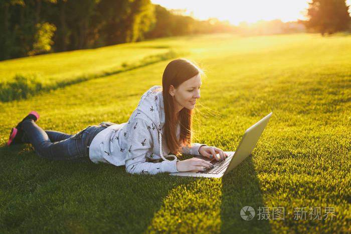 年轻聪明的女学生穿休闲服装。 女人躺在草地上，在户外绿草阳光草坪上的城市公园里，在笔记本电脑电脑上工作。 移动办公室。 自由职业