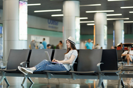 年轻冷静的旅行者旅游妇女坐在笔记本电脑上工作，而在国际机场的大堂等候。 周末出国旅行的乘客下车。 航空旅行飞行旅程概念