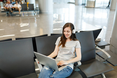 年轻冷静的旅行者旅游妇女带着耳机，听着音乐，在笔记本电脑上工作，等待在国际机场大厅。 周末出国旅行的乘客下车。 空中飞行概念