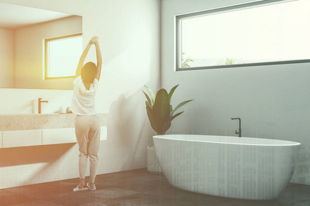 豪华浴室角落，白色墙壁，瓷砖黑色地板，白色浴缸和水槽。 狭窄的水平窗户。 斯堪的纳维亚风格。 穿睡衣的女人3D渲染色调图像双曝光