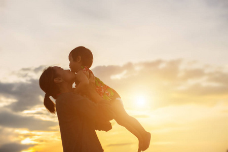 一个快乐的年轻女孩的剪影，他慈爱的母亲的手臂为一个拥抱，在日落前的天空中的一个夏天。