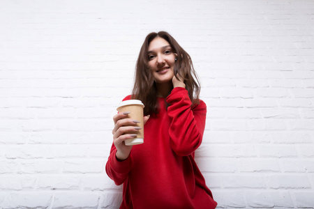 一个年轻漂亮的微笑女孩穿着一件红色毛衣，背景是现代白色砖块阁楼风格的墙，手里拿着一杯外卖咖啡，在电话里交谈。她看着观众。复制空间